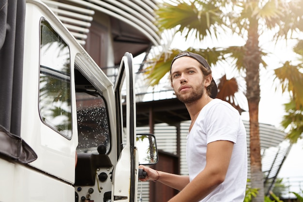 Fashionable young man with stylish beard wearing white t-shirt and baseball cap backwards looking away with confident and proud face expression while getting in his four-wheel drive vehicle