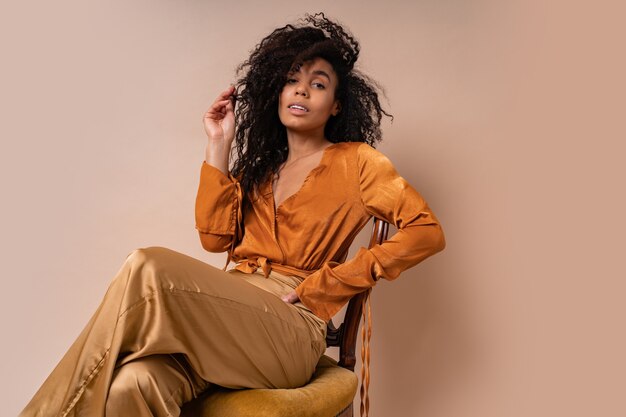 우아한 오렌지 블라우스와 빈티지 의자 베이지 벽에 앉아 실크 바지에 완벽한 곱슬 머리를 가진 유행 젊은 매혹적인 아프리카 모델.