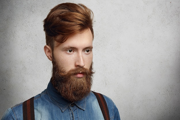 Fashionable young student with stylish haircut spending time indoors, standing against copy space wall for your promotional content.