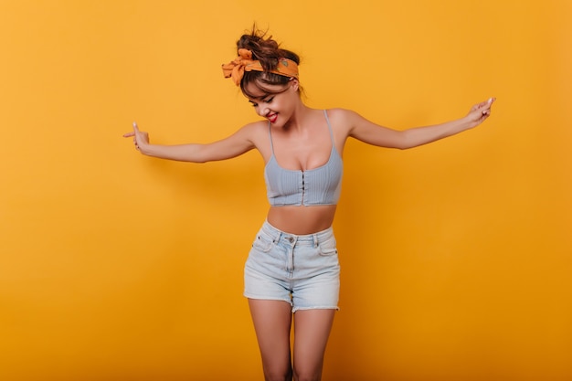 Fashionable young woman in cute tank-top posing on yellow space and waving hands