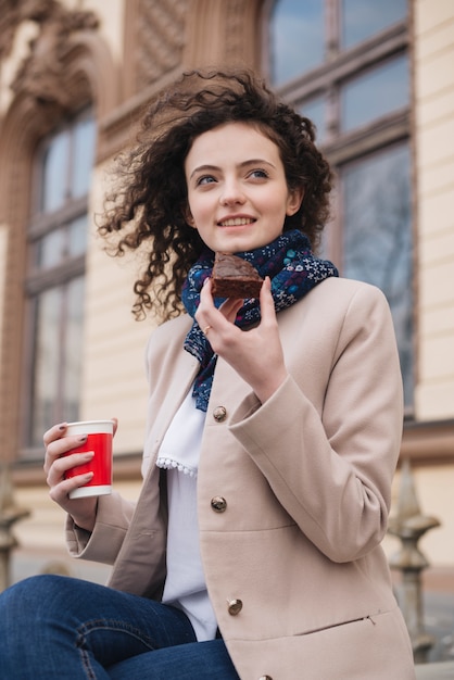 チョコレートケーキとコーヒーの使い捨てカップのスライスを楽しむファッショナブルな若い女性