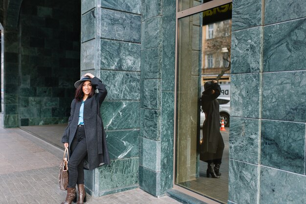 Fashionable young woman in grey coat, hat walking on street in city centre. Smiling, true emotions, stylish lifestyle, luxury clothes, elegant look.