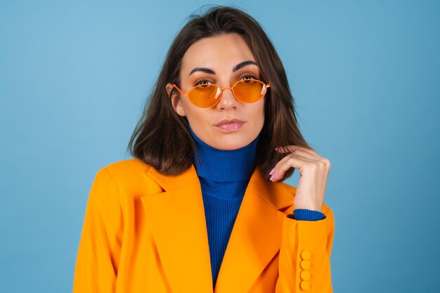 スタイリッシュな明るいオレンジ色のメガネのポーズで青い壁に膝丈と特大のジャケットのファッショナブルな若い女性