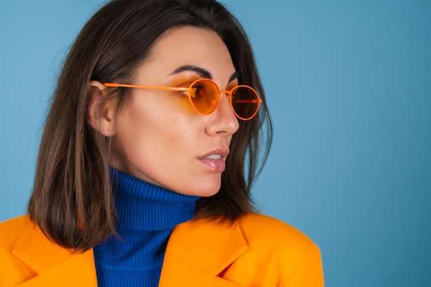 파란색 벽에 무릎 길이의 오버사이즈 재킷을 입은 세련된 젊은 여성이 세련된 밝은 오렌지색 안경을 쓰고 포즈를 취하고 있습니다.