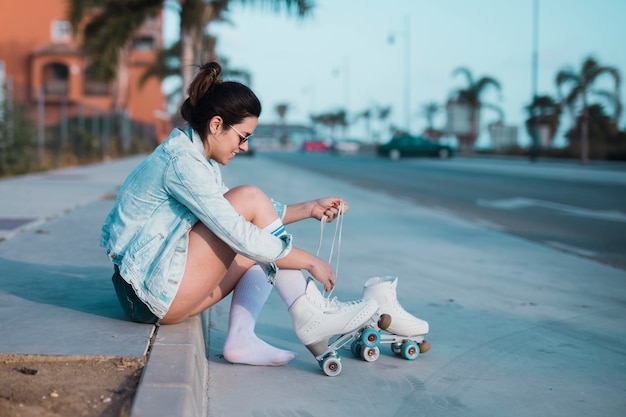 Free photo fashionable young woman sitting on sidewalk tying the lace of roller skate on street