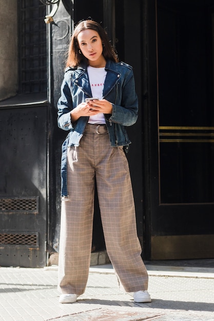 Free photo fashionable young woman standing on street holding mobile phone in hand