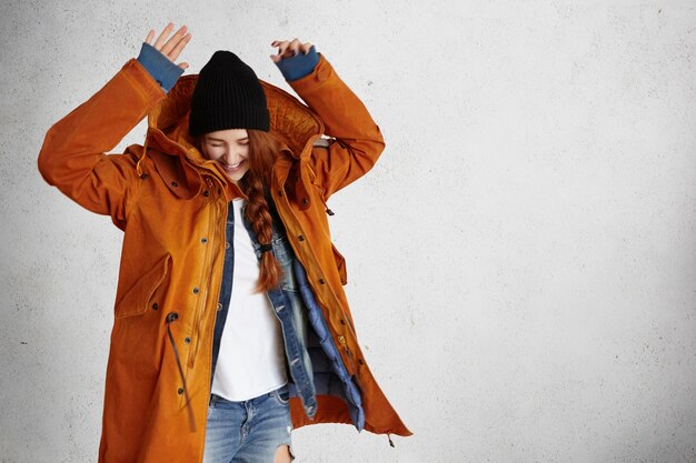 Fashionable young woman wearing red winter coat, black hat and ragged jeans raising hands in air