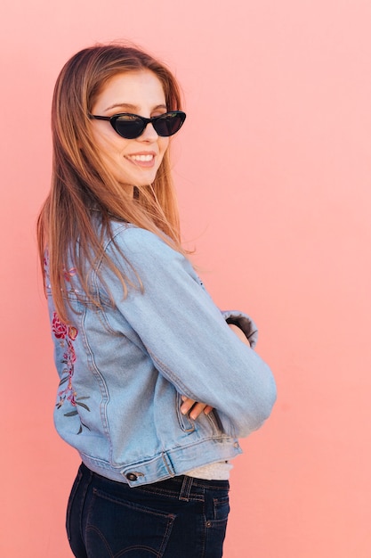 Foto gratuita occhiali da sole d'uso della giovane donna alla moda che esaminano spalla contro fondo rosa