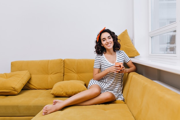 モダンなアパートメントのお茶のカップが付いているソファーでゾッとカットのカーリーブルネットの髪を持つファッショナブルな若い女性。居心地の良い時間、快適さ、自宅で、笑顔、陽気な気分