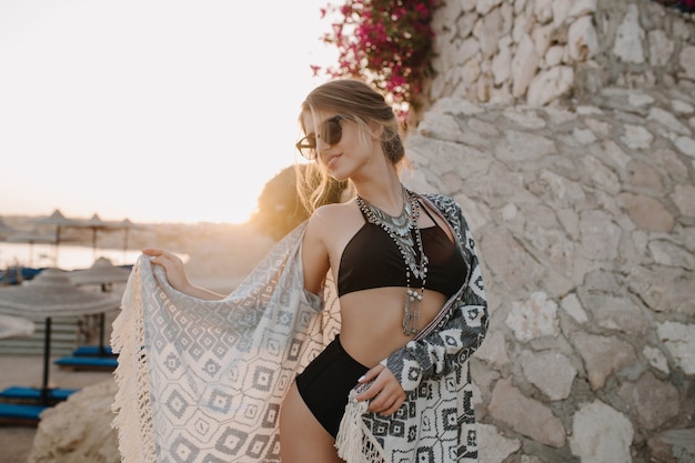 Модная модель позирует на пляже, девушка в сексуальном черном бикини, купальнике с завышенной талией, кардигане, накидке с орнаментом, красивый пляж, цветы, рок.