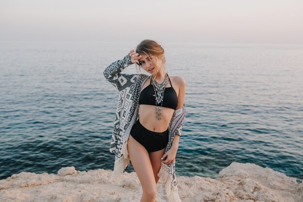 海、海の近くの夕日、ロックビーチでポーズをとっておしゃれなモデル。黒のビキニ、ハイウエストの水着、カーディガン、装飾品のケープを着ているセクシーな女性。