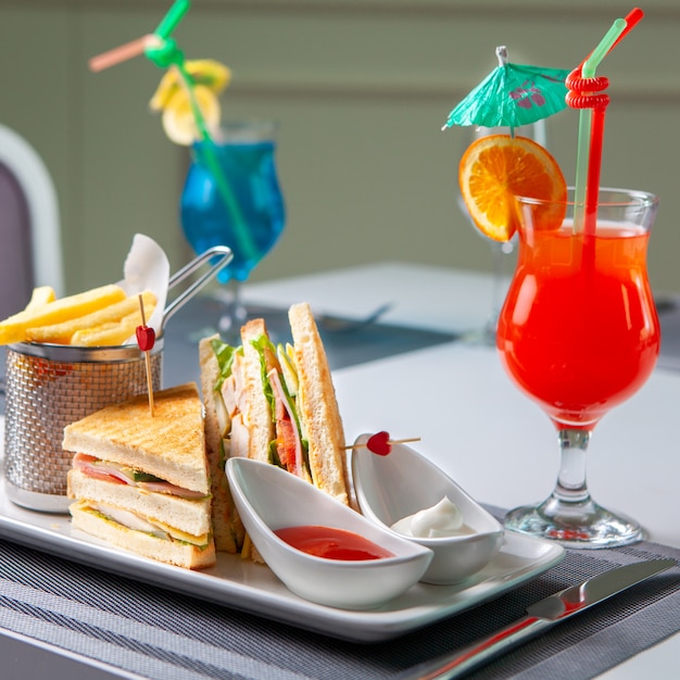 Fast food with sandwich, french fries, red cocktail, fork and knife on table, side view.