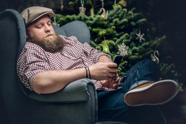 Толстый бородатый мужчина сидит на стуле и использует смартфон на фоне рождественской елки.