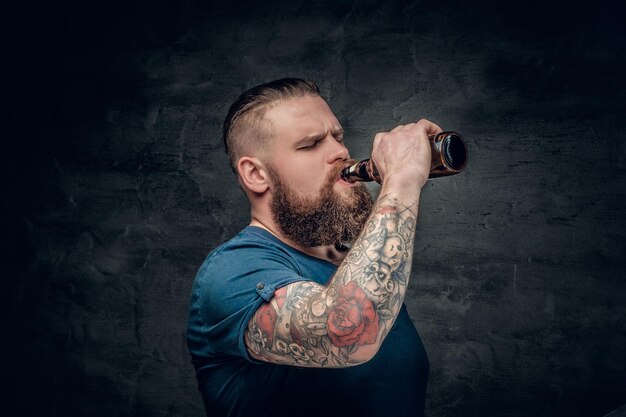 Fat bearded men with tattoos on arm drinks beer from a bottle.