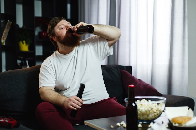 Толстяк пьет пиво, лежащее на диване