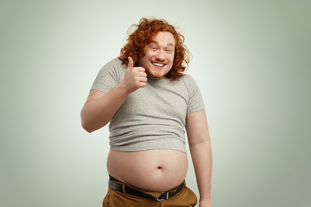 Толстый избыточный вес, рыжий кавказский мужчина в маленькой футболке показывает грудь и улыбается