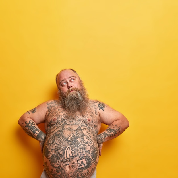 Free photo fat thoughtful man keeps hands akimbo, has big naked tattooed belly, thick beard, looks pensively upwards, has serious expression, thinks how to loose weight, isolated on yellow wall