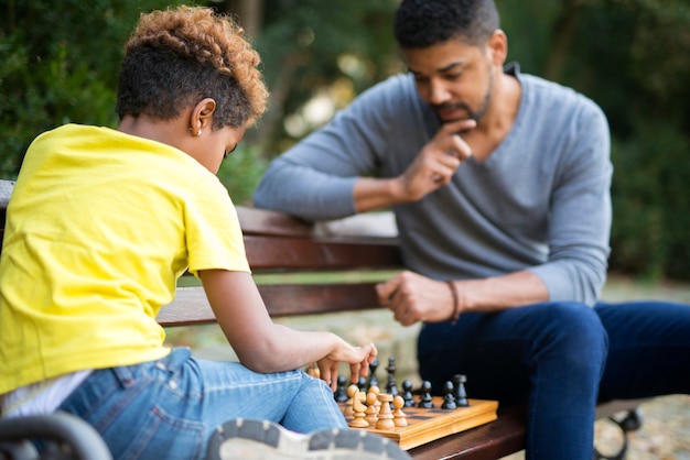 Отец и дочь играют в шахматы на скамейке в городском парке