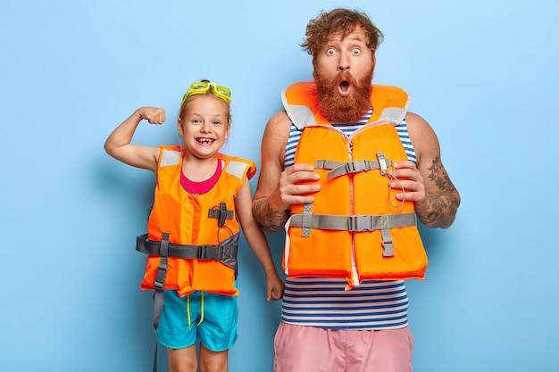 Foto gratuita padre e figlia in giubbotti di salvataggio di sicurezza avranno attività estreme sull'acqua. l'uomo barbuto scioccato dello zenzero guarda la telecamera