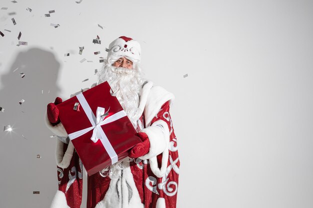 Дед Мороз в длинном теплом пальто, красных варежках и шляпе держит рождественский подарок с множеством конфеттов ...