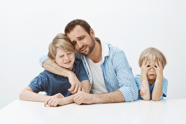 Father grief with sons while sitting at table, hugging boy and crying, being upset and unhappy while younger son having nothing to do
