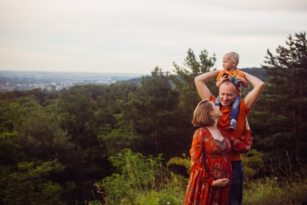 Father holds son in his arms posing with pregnant woman in the forest 