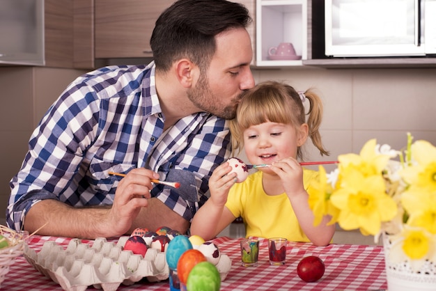 Отец целует голову своего маленького ребенка и рисует пасхальные яйца