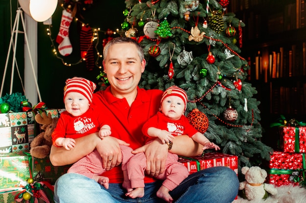 Father sits with twins in red suits before a Christmas tree 