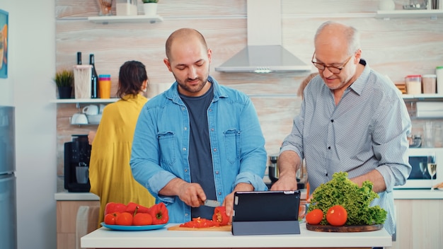 Padre e figlio che cucinano le verdure per cena usando la ricetta online sul computer del pc nella cucina di casa. uomini che usano la tavoletta digitale durante la preparazione del pasto. fine settimana rilassante e accogliente per famiglie allargate.