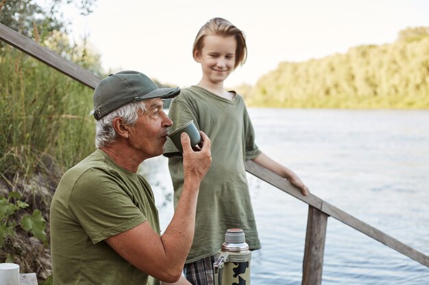 Father and son fishing at bank of river or lake, senior man drinking tea from thermos, family posing on wooden stairs leading to water, rest on beautiful nature.