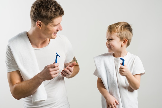 Father and son holding razor in hand looking at each other against white background