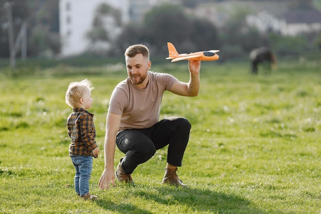 아버지와 아들은 비행기 장난감을 가지고 노는 여름 공원 야외에서 재미 청바지와 격자 무늬 셔츠를 입고 곱슬 유아 소년