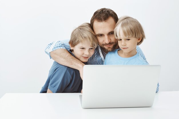 Father and sons talking with mom via video chat in laptop. Portrait of beautiful happy dad and boys hugging and gazing at notebook screen, watching touching videos or cute photos