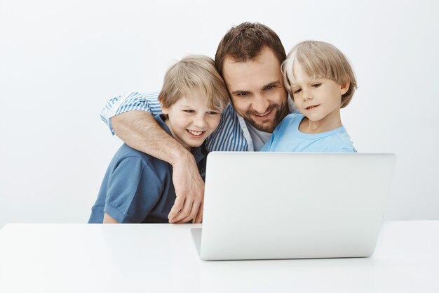 Father talking to wife hugging charming sons. Portrait of happy carefree blond dad cuddling boys and smiling broadly at laptop screen while talking with mom or watching touching photos together