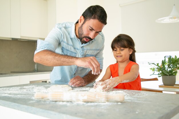빵이나 파이를 굽는 그의 소녀를 가르치는 아버지. 집중된 아빠와 딸 밀가루 지저분한 식탁에 반죽을 반죽. 가족 요리 개념