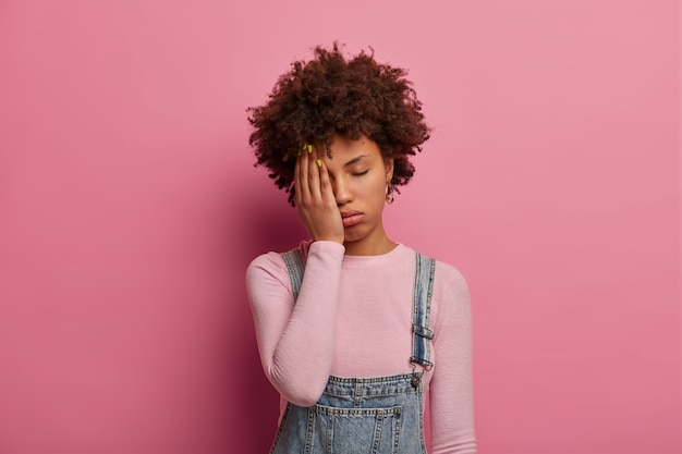 Fatigue curly haired woman feels bored and distressed, wants to sleep, covers half of face with palm, keeps eyes shut, wears fashionable clothes, poses against pink wall. Tiredness concept