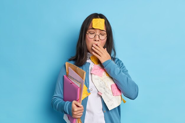 Fatigue exhausted female student yawns and has sleepy expression worked long hours covers mouth with hand carries folders with papers tries to learn all material for exam.