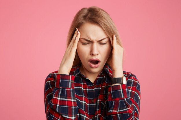 Fatigue stressful young female model keeps hands on head, closes eyes and opens mouth in desperation