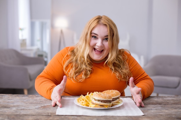 가장 좋아하는 음식. 테이블에 앉아 감자 튀김과 샌드위치를 먹는 쾌활한 과체중 여성