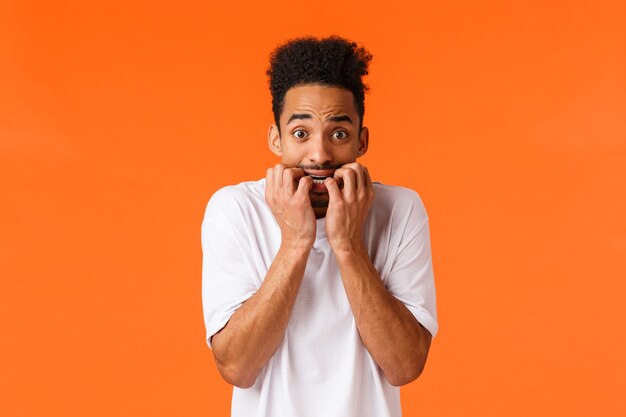 Fear, horror movies, emotions concept. Frightened and insecure, young timid hipster african american male, biting fingernails, staring camera anxious, scared someone know, orange background