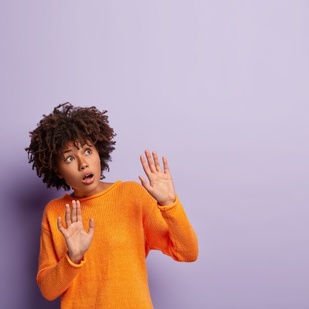 Fearful dark skinned female keeps palms forward, tries to defense herself, focused above, afraids of something terrible, wears orange jumper, isolated against purple wall. Oh no, something is falling