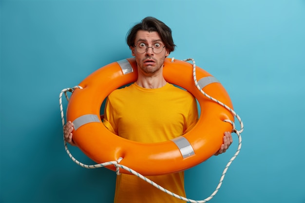 Fearful man afraids of swimming in deep sea, poses with inflated lifebuoy, listens advice of instructor, wears spectacles and glasses, poses 