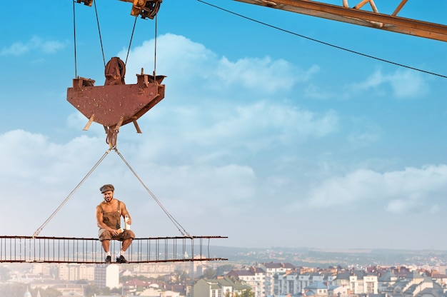 Fearless man. Handsome cheerful hunky builder enjoying lunch sitting on a crossbar hanging on a crane at the construction