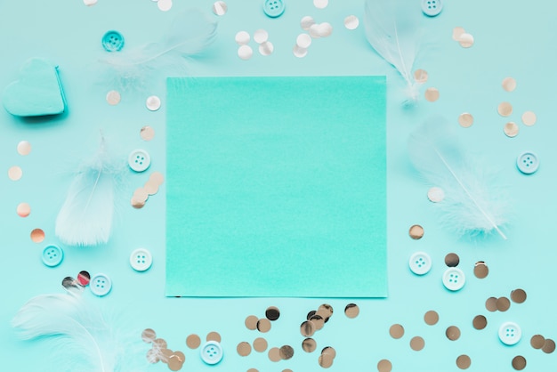 Feather; sequins; buttons surrounded around the turquoise paper on teal backdrop