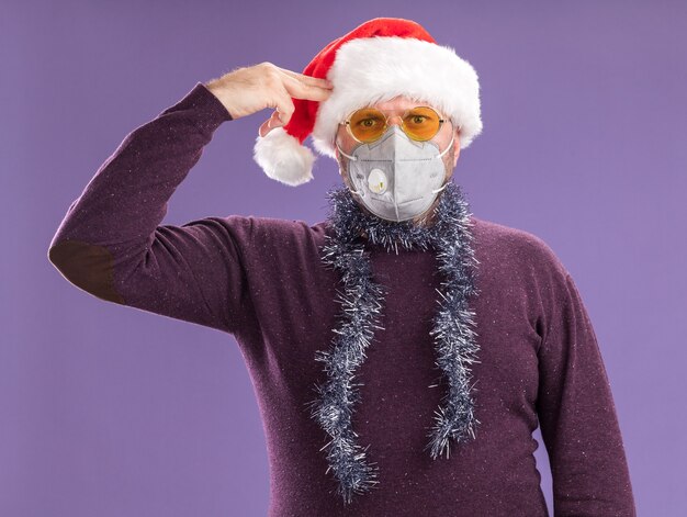 Fed up middle-aged man wearing santa hat and protective mask with tinsel garland around neck with glasses  doing suicide gesture isolated on purple wall