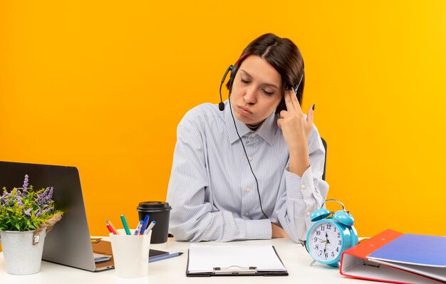 Fed up young call center girl wearing headset sitting at desk doing pistol gesture with hand gesturing suicide with closed eyes isolated on orange 