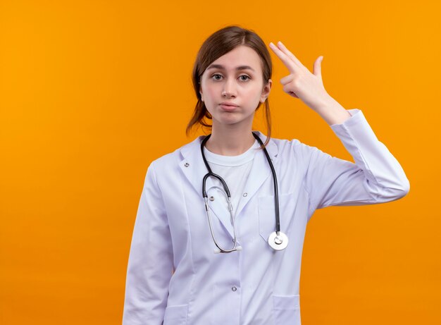 Fed up young female doctor wearing medical robe and stethoscope doing suicide gesture on isolated orange space with copy space
