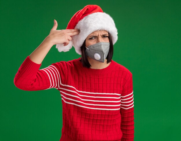 Fed up young girl wearing santa hat and protective mask  doing suicide gesture isolated on green wall