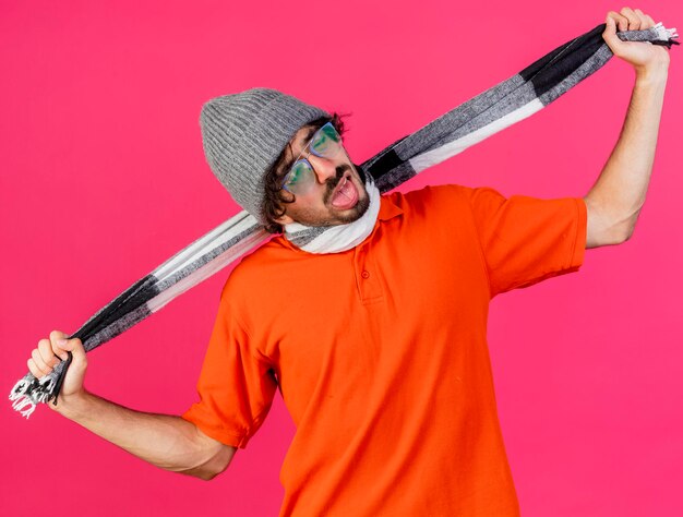 Fed up young ill man wearing glasses winter hat and scarf choking himself with scarf isolated on pink wall