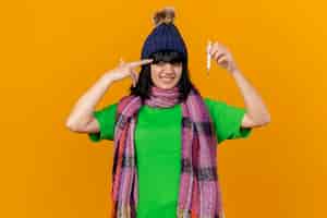 Foto gratuita stufo di giovane donna malata che indossa cappello invernale e sciarpa tenendo il termometro facendo gesto di suicidio isolato sulla parete arancione
